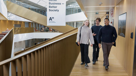 Three students walk in a bright wooden hallway with a large Better Business Better Society banner in the background