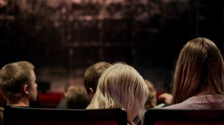 Yleisöä selin punaisilla teatteripenkeillä hämärää näyttämöä kohti