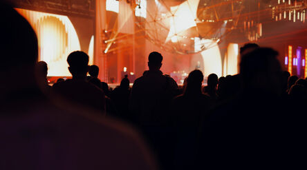 Crowd watching stage show in warm orange light, with abstract geometric structures above