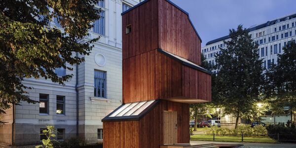 A modern, two-storey wooden house with large windows, situated in a courtyard with trees and buildings in the background.