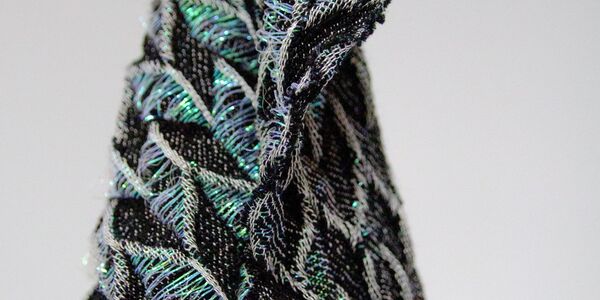 Close-up of a textured, triangular fabric with black, silver, and iridescent green threads, resembling a folded pattern.
