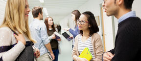 Several students, women and men discussing in the school hallway. One of the women is holding a yellow book.