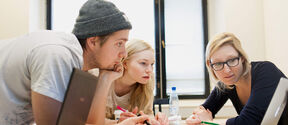 A group of three students engaging in teamwork, one presenting something from her laptop's screen