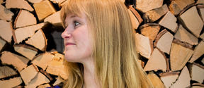 Anniina Suominen, an Associate Professor of Art Pedagogy at Aalto University’s School of Arts, Design and Architecture. She is standing in front of the camera, smiling, arms crossed. In the background there is a stack of logs.