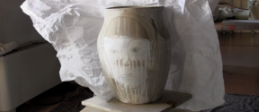 a ceramic vase with a face painted on it. behind a wrinkled white paper