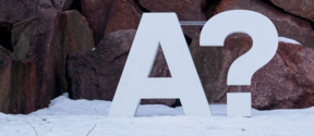 Iso valkoinen kirjain 'A' ja kysymysmerkki seisovat lumessa suurten kivien taustalla.