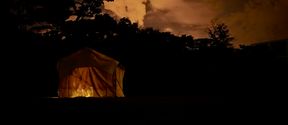 Still from the film Like Shadows Through Leaves, 2021. A lamp-lit tent stands in a dark landscape, it's orange interior matching the orange skyline