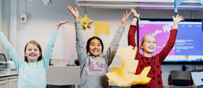 Three children with arms raised in a classroom, surrounded by colourful paper crafts and a monitor in the background.
