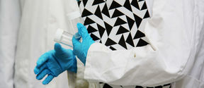A person with a lab coat and protective gloves carrying test tubes and a black-and-white notebook. Their face is not pictured.