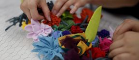 Lähikuva käsistä, jotka kiinnittävät kankaasta tehtyjä kukkia kanaverkkoon