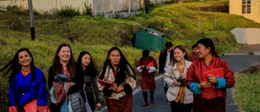 Nine students wearing bright colours carrying notebooks walking down a street with buildings in the background