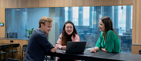 Two students and a professor sitting around a table, talking and looking at laptop screen.
