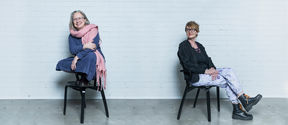 Laura Arpiainen ja Susanna Helke istumassa mustilla tuoleilla valkoisen tiiliseinän edessä. Arpiainen istuu tuolin selkänojan päällä jalat istuinosalla ja Helke katsoo häntä hymyillen.