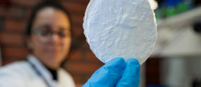 Colour photo of a blurry person in a white lab coat holding an in-focus white paper-like wafer in their blue gloved hand