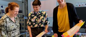 A teacher and two researchers smiling and sorting through paper presentations in a classroom