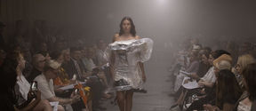 Catwalkilla kävelevä malli esittelee muotisuunnittelijan luomaa asua, katsojat istuvat katsomassa näytöstä