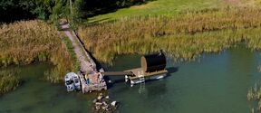 Aerial view of a seaside pier with a floating sauna, small boat docked nearby, and grassy shore with trees.