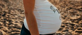 A close-up of a pregnant person wearing a white T-shirt, standing on a sandy beach at sunset.