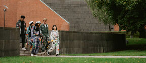 A group of people wearing colourful student overalls walk down steps beside a brick building and a large tree.