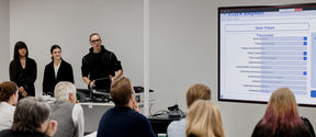 Three people are presenting to an audience, with a large screen displaying a form on the right.