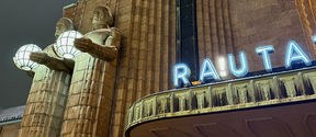 Närbild av Helsingfors centralstation med två stenstatyer som håller sfäriska lampor och skylten 'RAUTA'.