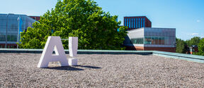 A large white 'A!' sculpture on the rooftop of the Undergraduate centre. A large tree and other buildings in the background.