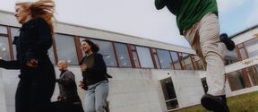 Fyra personer springer på en gräsyta nära en modern byggnad med stora fönster.