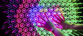 Two hands hover over a board of colourful circular LED lights and switches arranged in a pattern.