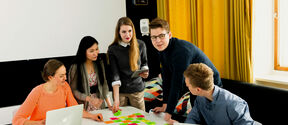 A group of people collaborate around a table covered with sticky notes and laptops in a modern office.