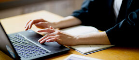 Person typing on a laptop with a notebook and tablet on the desk.
