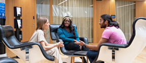Three people sit in white chairs around a table with a laptop in a modern office space with wooden walls.