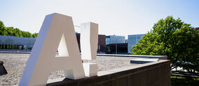 Suuri valkoinen 'A!'-logo katolla, taustalla modernit rakennukset ja vihreät puut.