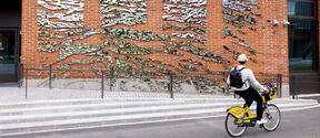A person rides a yellow bike past a brick wall with abstract metal art installations. The rider wears a hat and has a backpack.