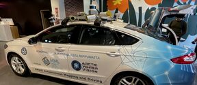 A white and blue autonomous car with various sensors on the roof, parked indoors with its boot open.