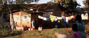 A rural scene with people washing clothes outside a small house. Colourful laundry hangs on a line. Trees and grass are visible.