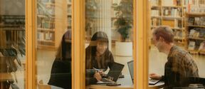 Kolme henkilöä työskentelee kannettavien tietokoneiden parissa pyöreän pöydän ääressä kirjastossa, lasiseinien läpi.
