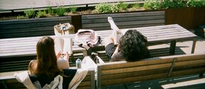 Two people are sitting on wooden benches outside, reading books. There is a grassy area and a bike park in the background.