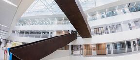Modern atrium in an office building with two dark metal diagonal beams, large windows, and people wearing safety gear.