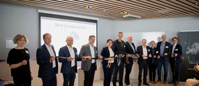 Group of people standing in a room holding a ribbon, with presentation screen and institute banner in the background.