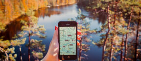 Person holding smartphone with map app in front of scenic lake view surrounded by forest in autumn.