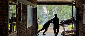 Two people push a stretcher through a brick-walled corridor towards the outdoors. Trees and a parked car are visible.