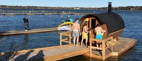 Four people stand by a floating sauna on a sunny day near the shore. The sea and distant land are visible in the background.