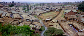 A view over densely packed houses in a slum area. Rusty metal roofs, scattered vegetation, and a narrow stream are visible.