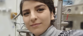 Person wearing a patterned knit sweater and grey turtleneck in a science laboratory with metal equipment in the background.