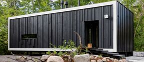 Modern black wooden house with a single narrow window, surrounded by rocks and plants, nestled in a leafy forest area.