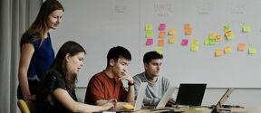 Four people collaborate at a table with laptops. Sticky notes are on a whiteboard behind them, showing various colours.