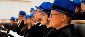 People wearing blue academic hats and black gowns seated in rows during a ceremony.
