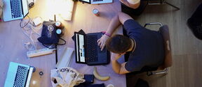 People working on laptops at a cluttered table with drinks, cables, and bananas.
