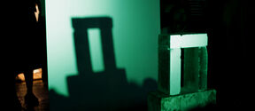 A sculpted model casting a shadow resembling Stonehenge onto a green-lit screen, with people standing nearby.