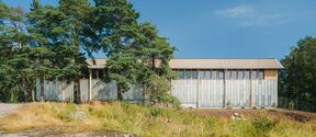 Modern storage building with grey facade with wooden details and windows surrounded by trees and grass under a clear blue sky.